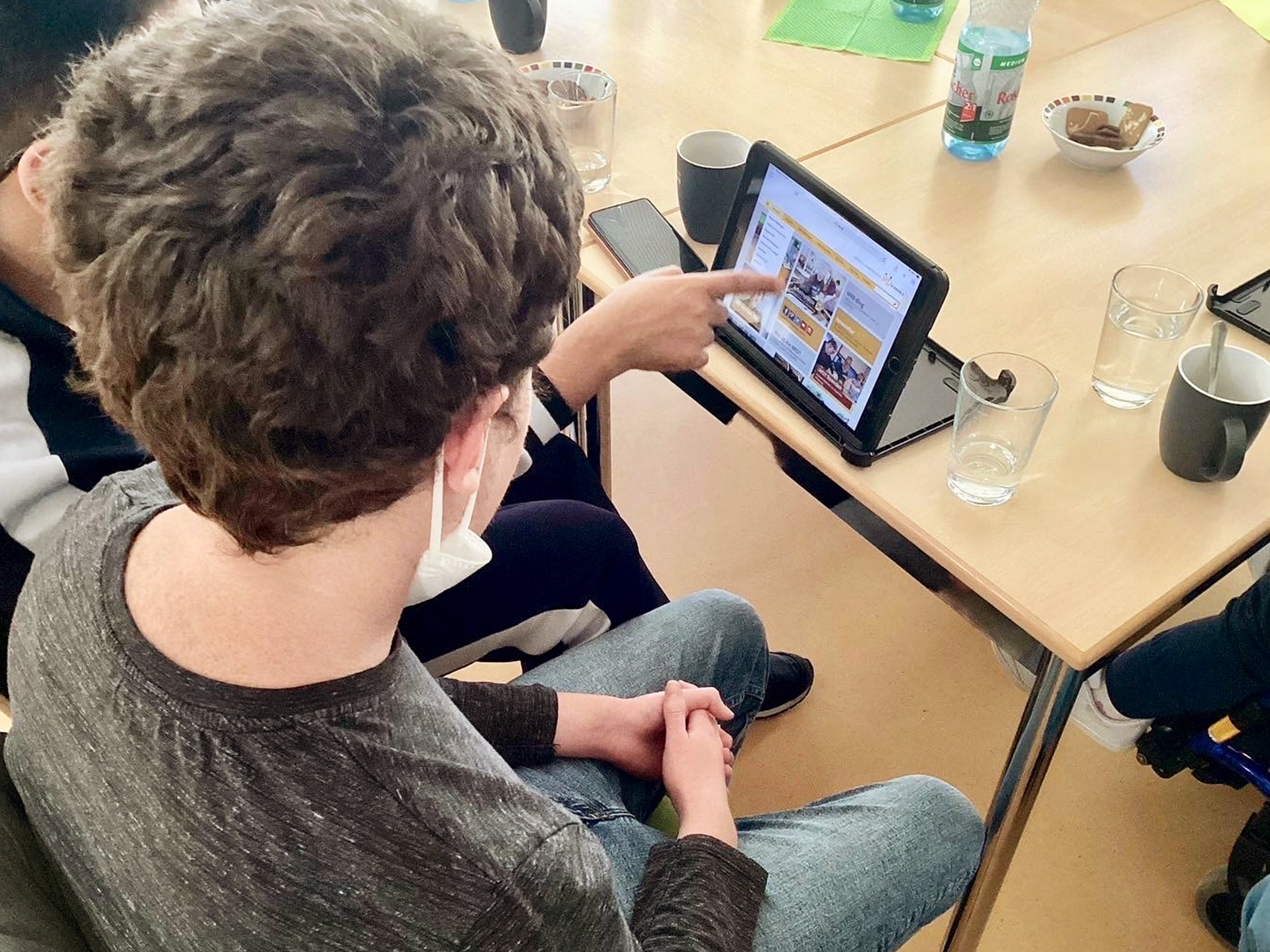 2 Lernende in einem Workshop sitzen an einem Tisch. Vor Ihnen ist ein iPad. Einer der beiden zeigt auf das iPad. Auf dem iPad ist eine Webseite zu sehen.