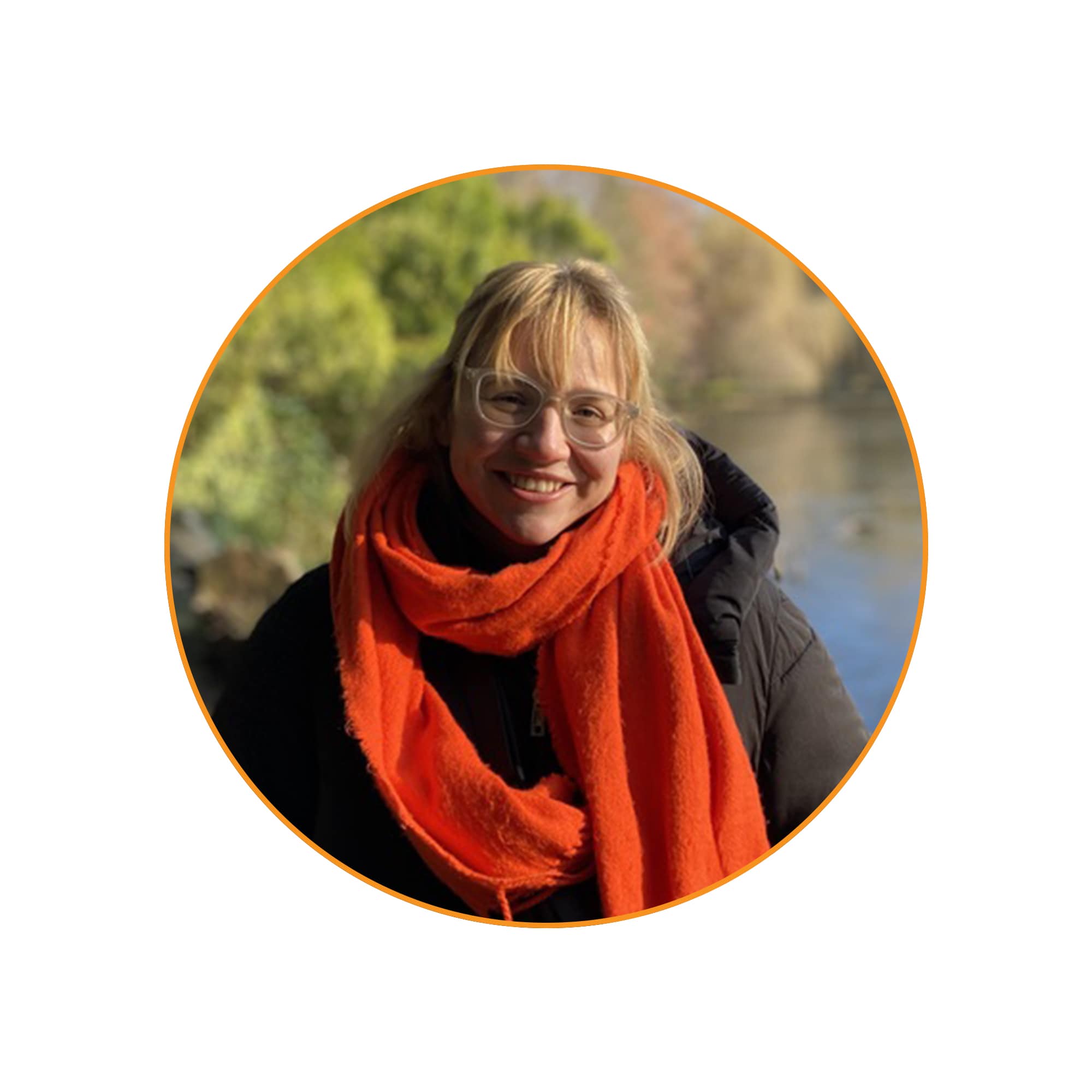 Profilbild Veronika Meister: Eine lächelnde Frau mit blonden Haaren und Brille steht vor einem Gewässer inmitten einer natürlichen Umgebung. Sie trägt eine schwarze Winterjacke und einen leuchtend orangefarbenen Schal. Die Sonne scheint, wodurch ihr Gesicht und die Umgebung warm beleuchtet werden. Im Hintergrund sind grüne Bäume, herbstlich gefärbte Sträucher und das ruhige Wasser eines Sees oder Flusses zu sehen.