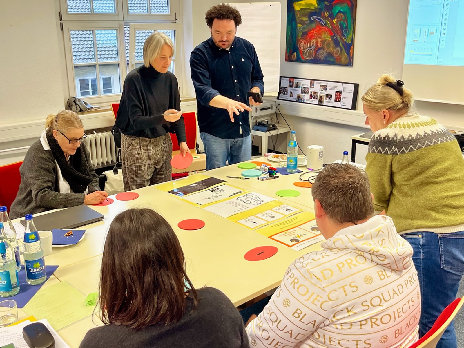 Mehrere Personen sitzen und stehen um einen Tisch in einem Workshop-Raum während eines Brainstorming Prozesses. Auf dem Tisch liegen verschiedene Materialien, darunter große Papierbögen mit Diagrammen, bunte Moderationskarten, Marker und Wasserflaschen. Eine Frau in schwarzem Pullover hält eine rote Karte, während ein Mann mit dunkler Lockenfrisur in einem blauen Hemd gestikulierend etwas erklärt. Eine Leinwand im Hintergrund zeigt eine Präsentation, daneben hängt ein buntes abstraktes Gemälde an der Wand. Die Teilnehmenden wirken konzentriert und engagiert.