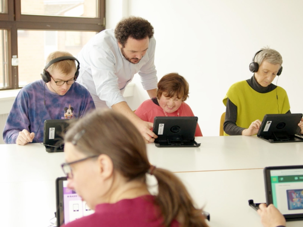 Mehrere erwachsene Menschen mit und ohne Behinderung sitzen mit Kopfhörern und Tablets an einem Tisch und arbeiten konzentriert an einem digitalen Lernmodul. Eine Person unterstützt die Gruppe und beugt sich zu einer Teilnehmerin, um Hilfe zu leisten. Im Vordergrund sind zwei weitere Personen mit Tablets zu sehen, auf deren Bildschirmen die Inhalte des WEIDI-Lernmoduls sichtbar sind.