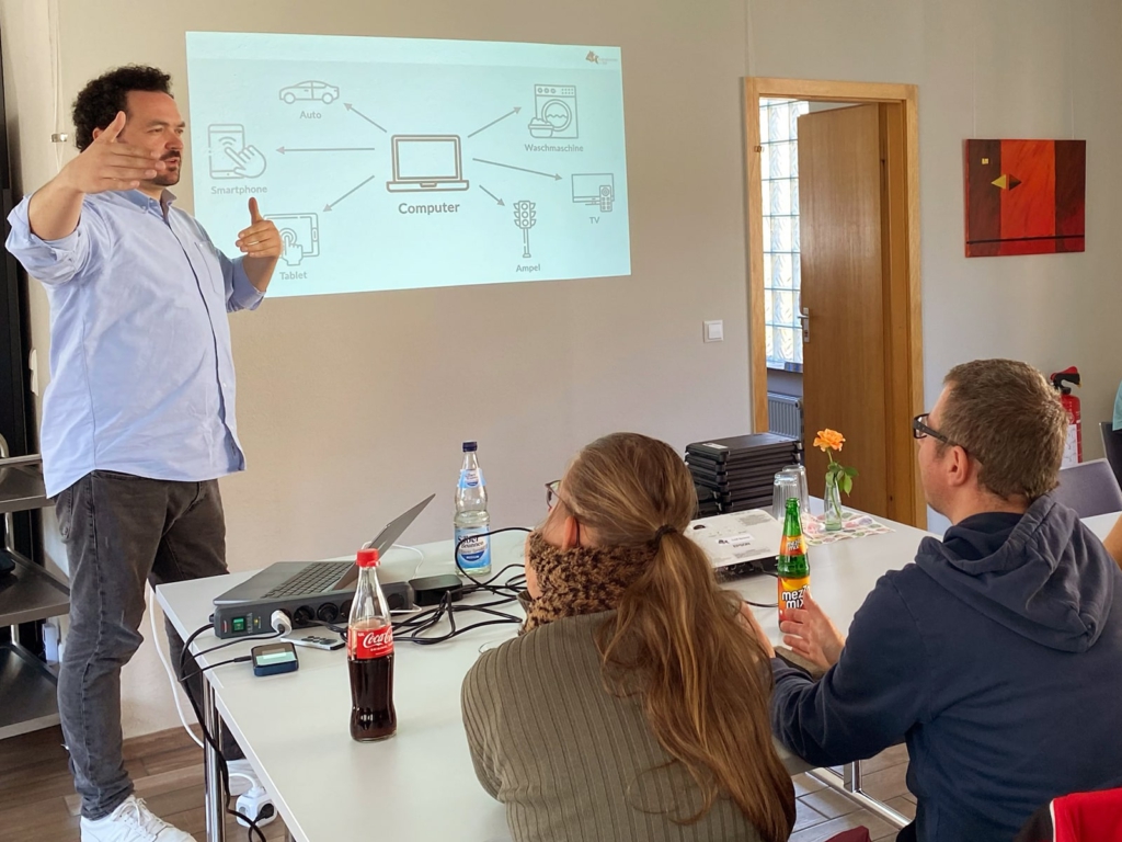 Workshops Einfach Computer -min Das Bild zeigt eine Person, die vor einer kleinen Gruppe von Menschen mit und ohne Behinderung in einem Seminarraum steht und mit Gestik einen Vortrag hält. Auf der Wand ist eine Folie projiziert, die einen Computer in der Mitte zeigt, von dem Pfeile zu Symbolen für Smartphone, Tablet, Auto, Waschmaschine, TV und Ampel führen.