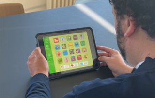in Mann mit dunklem, lockigem Haar und dunkelblauer Jacke sitzt an einem Tisch und hält ein Tablet in den Händen. Auf dem Bildschirm ist eine App mit großen, farbigen Symbolen und Icons zu sehen, die an eine leicht verständliche Benutzeroberfläche erinnert. Im Hintergrund stehen bunte Stühle und eine weiße Heizung.