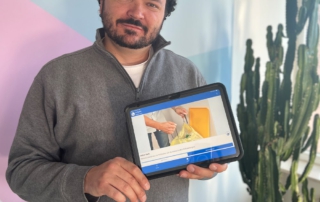Mann mit dunklen, lockigen Haaren und Bart hält ein Tablet in beiden Händen. Auf dem Tablet ist die InA.Coach-App geöffnet, die ein Bild von Mülltrennung zeigt. Der Mann trägt ein graues Sweatshirt und steht vor einer Wand mit pastellfarbenen Flächen und einer großen Zimmerpflanze (Kaktus).