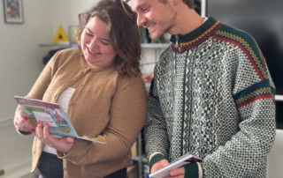 Zwei junge Erwachsene stehen nebeneinander in einem Raum und schauen gemeinsam in eine bunte Broschüre in Leichter Sprache. Die Person links trägt eine braune Strickjacke und lächelt, während sie das Heft hält. Die Person rechts trägt einen grünen Norweger-Pullover mit bunten Mustern und hält ebenfalls ein Heft in der Hand. Im Hintergrund sind ein Regal, Pflanzen und ein großer Bildschirm zu sehen.