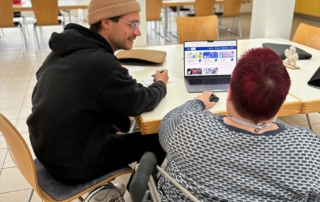 Ein junger Mann mit Brille, schwarzem Hoodie und beigefarbener Mütze sitzt an einem Tisch neben einer Person im Rollstuhl. Beide schauen auf einen Laptop, auf dem die Webseite „Medien leicht erklärt“ geöffnet ist. Sie befinden sich in einem hellen Aufenthaltsraum mit Holzstühlen und großen Fenstern.