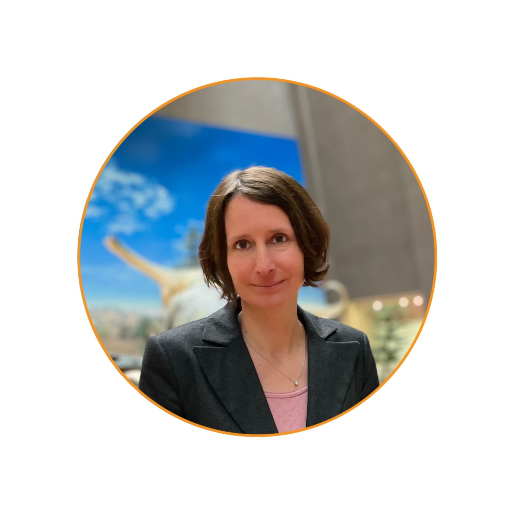 Portraitfoto einer Frau mit braunen, kinnlangen Haaren in einem dunklen Blazer vor einem unscharfen Hintergrund mit einem Dinosaurier-Modell und blauer Himmelsszene.