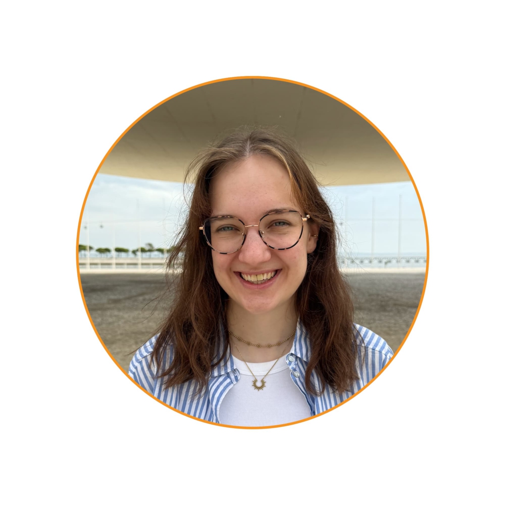 Portrait: Junge Frau mit langen braunen Haaren, Brille und blau-weiß gestreiftem Hemd steht lächelnd im Freien. Im Hintergrund sind ein gepflasterter Platz, ein modernes Dach und eine Brücke über dem Wasser zu sehen.