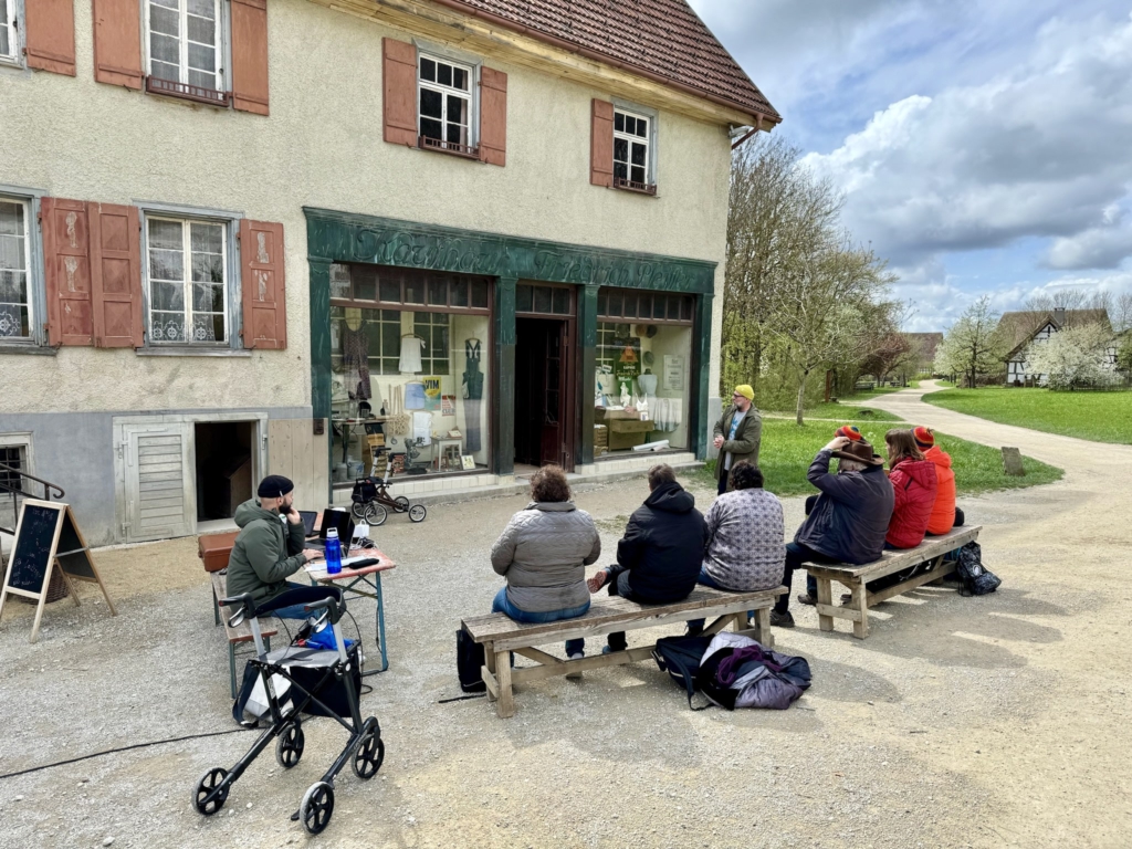 Foto: Eine Gruppe von Menschen mit und ohne Beeinträchtigungen sitzt auf zwei Holz­bänken vor einem historischen Kaufhaus mit beigen Außenwänden und roten Fensterläden. Vor dem Eingang steht eine Person und spricht zur Gruppe. Links sitzt eine weitere Person an einem kleinen Tisch mit Laptop. Ein Rollator steht im Vordergrund. Im Hintergrund führt ein Weg durch eine grüne Landschaft mit Bäumen und Fachwerkhäusern.