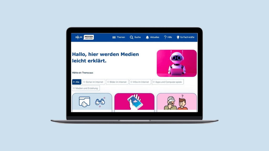 Foto: Ein geöffneter Laptop vor hellblauem Hintergrund zeigt die Startseite der Website „Medien leicht erklärt“. Auf dem Bildschirm steht groß „Hallo, hier werden Medien leicht erklärt“. Darunter befindet sich die Auswahl „Wähle ein Thema aus“. Die sichtbaren Filter sind „Alle“, „Sicher im Internet“, „Bilder im Internet“, „Infos im Internet“, „Apps und Computerspiele“ sowie „Medien und Erziehung“. Rechts im oberen Bereich des Inhalts wird ein Bild eines kleinen Roboters mit Laptop gezeigt. Unter dem Einführungsbereich sind mehrere farbige Kacheln mit unterschiedlichen Symbolen zu sehen, die verschiedene Medienthemen darstellen.
