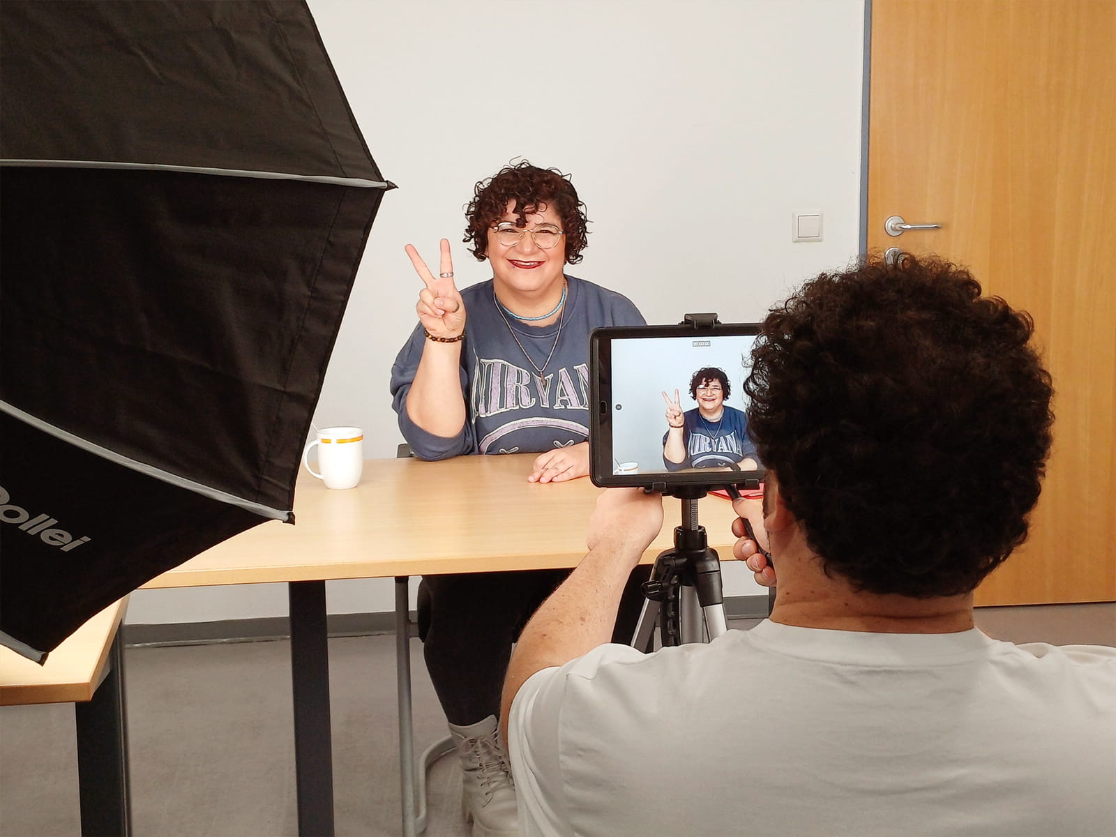 Foto: Eine Frau mit kurzen lockigen Haaren und Brille sitzt an einem Tisch und zeigt lächelnd ein Friedenszeichen in die Kamera. Vor ihr steht ein Tablet auf einem Stativ, dahinter sitzt eine Person mit kurzen Haaren und filmt. Seitlich steht eine Softbox Leuchte.