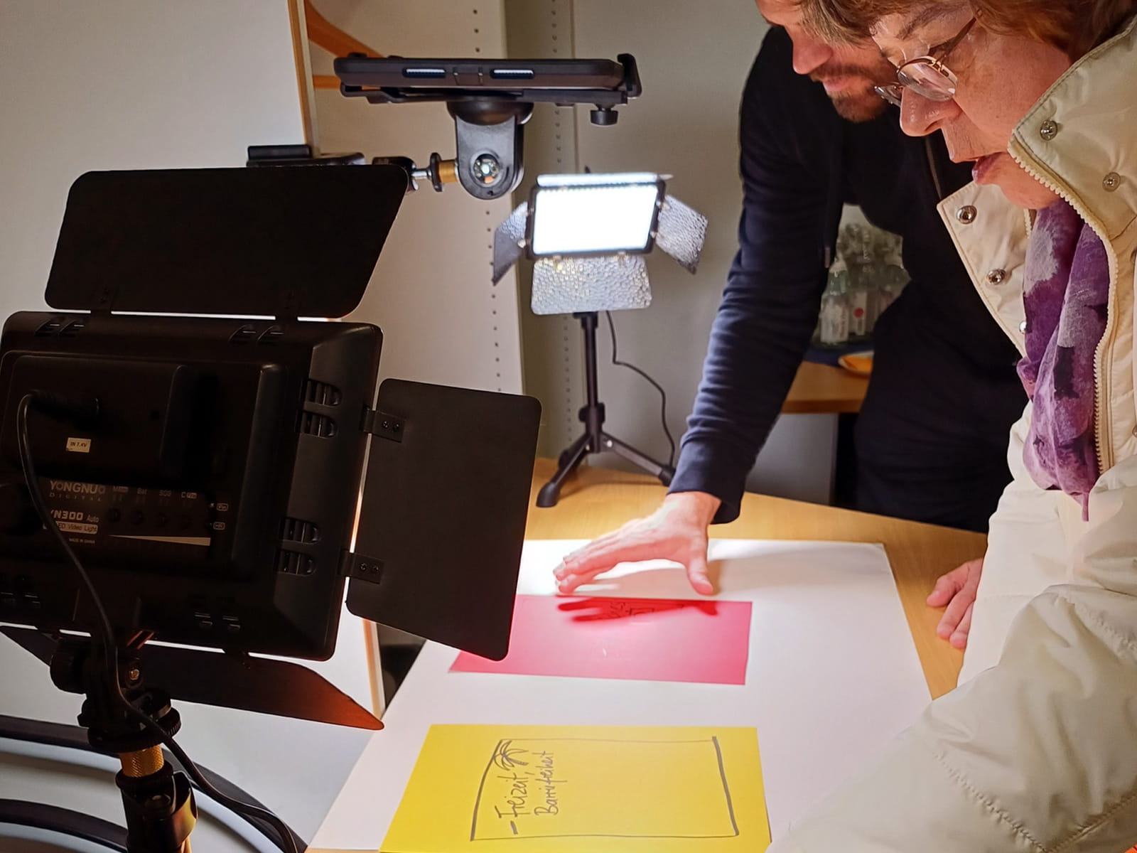 Foto: Eine ältere Frau mit Brille und heller Jacke und ein Mann mit Bart stehen an einem Tisch mit Videoleuchte und Kameraaufbau und betrachten ein Blatt Papier mit farbigem Hintergrund unter dem Licht.