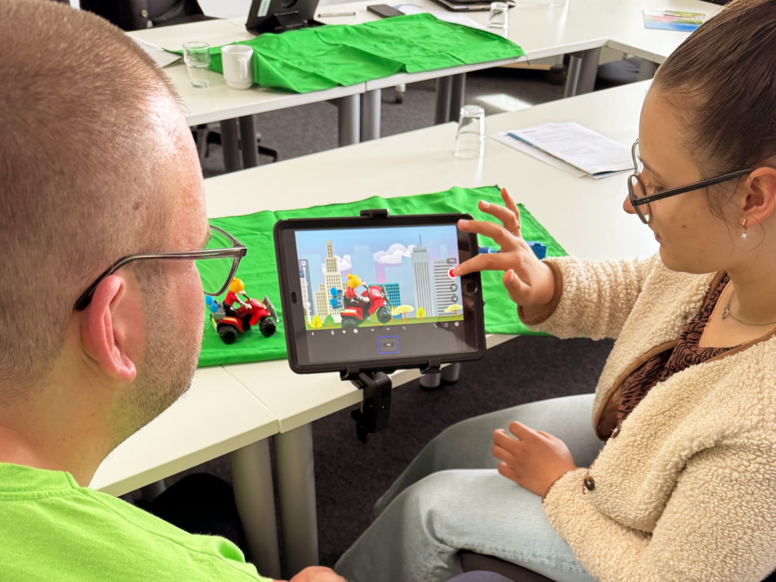 Foto: Eine junge Frau mit Brille und zusammengebundenen Haaren sitzt neben einem Mann mit kurz rasierten Haaren und Brille an einem Tisch und bedient ein Tablet auf einem Stativ. Auf dem Bildschirm ist eine Stop Motion Szene mit einer Spielfigur auf einem roten Quad vor einer gezeichneten Stadt zu sehen, daneben steht das passende Spielzeugfahrzeug auf grünem Stoff.