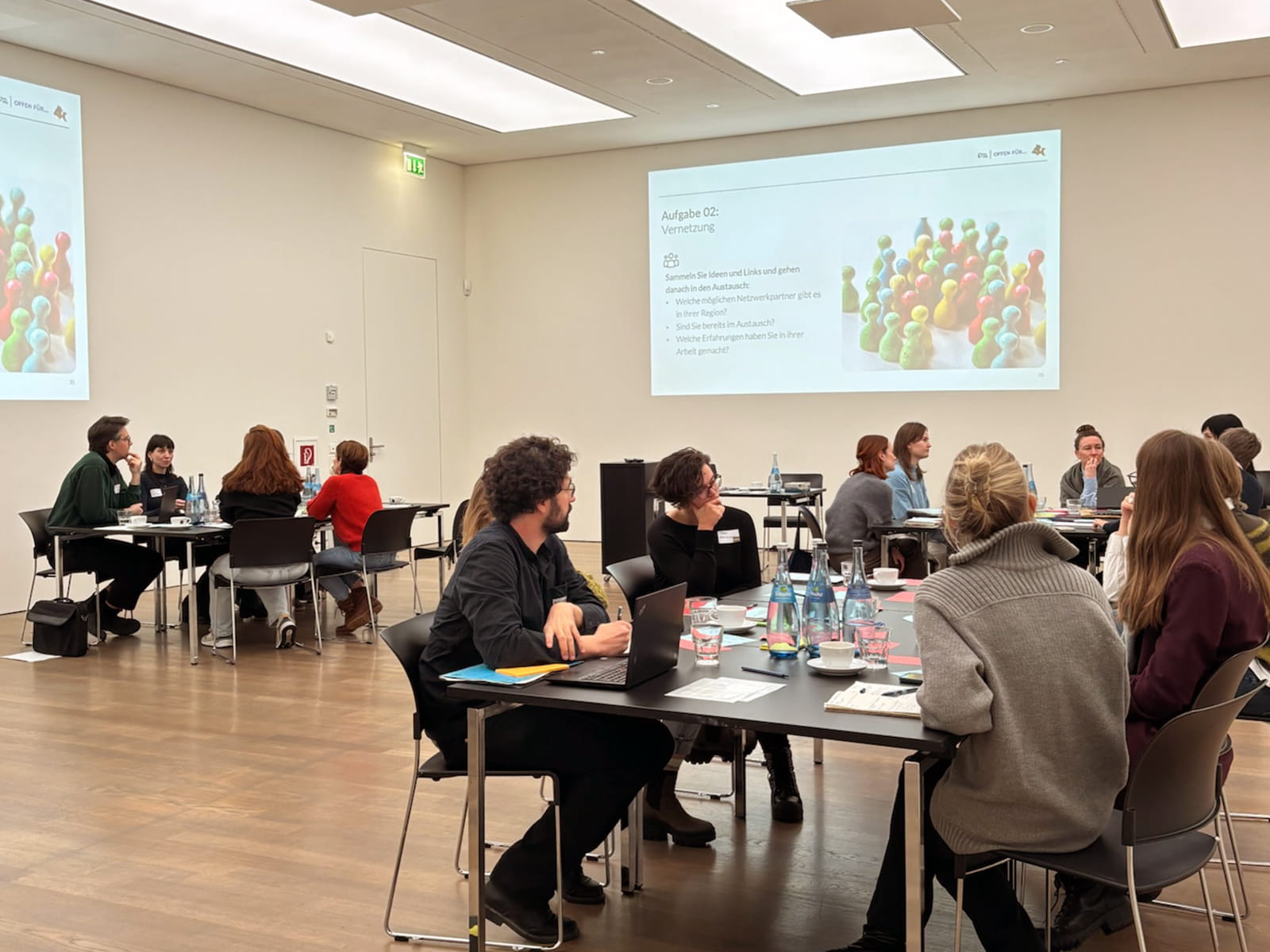 Coaching Kultur 01 Foto: Mehrere Arbeitsgruppen sitzen an Tischen in einem hellen Seminarraum und diskutieren gemeinsam. Auf den Tischen liegen Notizkarten, Laptops, Getränke und Arbeitsmaterialien. An der Wand ist eine Präsentationsfolie mit der Überschrift „Aufgabe 02: Vernetzung“ zu sehen. Die Teilnehmenden arbeiten in kleinen Gruppen und sprechen über mögliche Netzwerkpartner, Erfahrungen und Austauschmöglichkeiten.