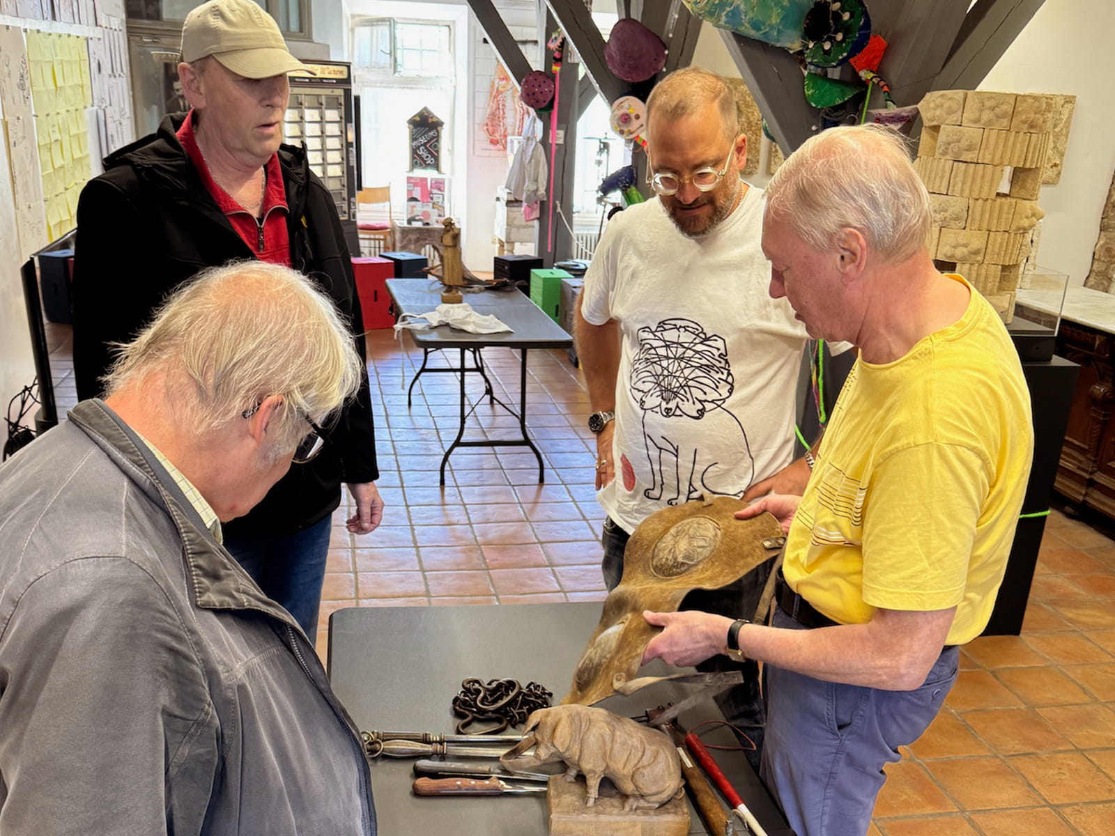 Coaching Kultur 02 Foto: Vier Personen stehen um einen Ausstellungstisch und betrachten historische Gegenstände. Drei Männer davon sind blind. Auf dem Tisch liegt auch ein Blindenstock. Ein älterer Mann in gelbem T-Shirt hält ein großes hölzernes Objekt in den Händen und zeigt es den anderen. Auf dem Tisch liegen verschiedene Werkzeuge, Ketten, Messer sowie eine geschnitzte Tierfigur. Die Teilnehmenden beobachten das Objekt aufmerksam und tauschen sich darüber aus.