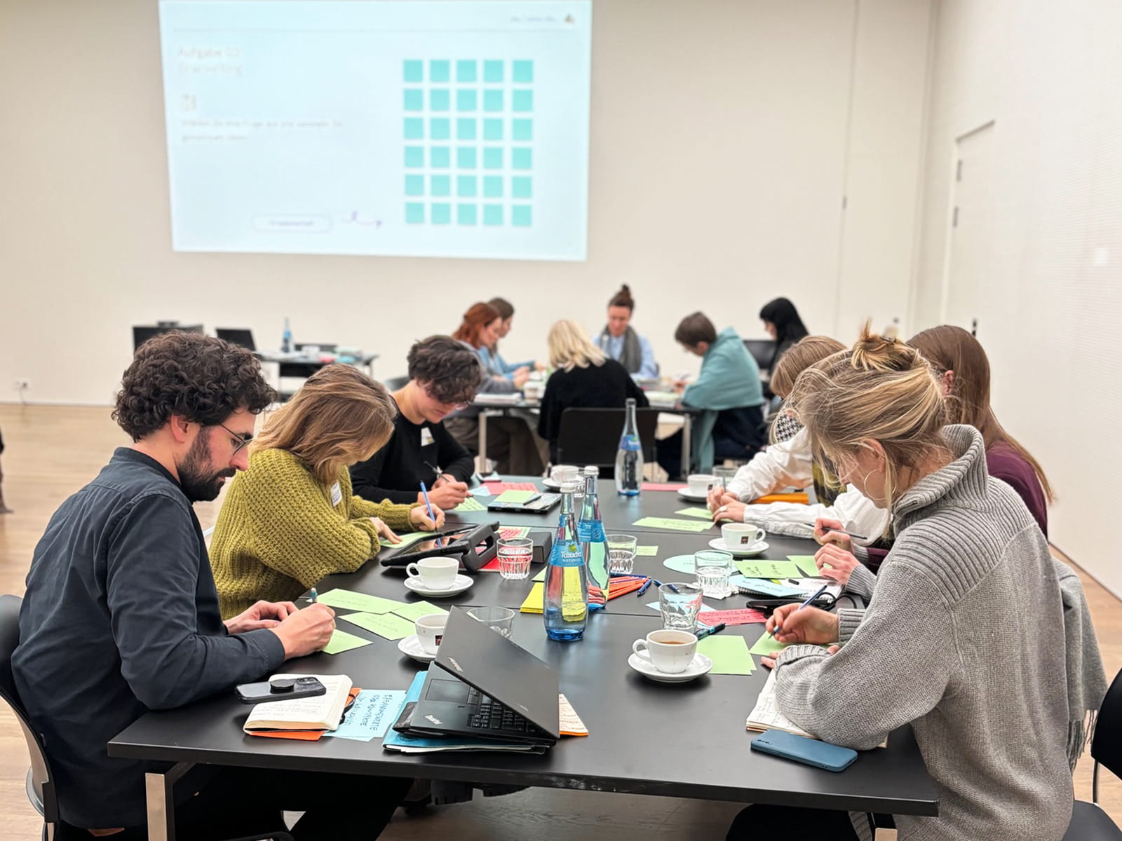 Fortbildung 01 Foto: Mehrere Personen sitzen an langen Tischen in einem hellen Seminarraum und schreiben auf farbige Moderationskarten. Auf den Tischen stehen Notizbücher, Laptops, Smartphones, Wasserflaschen, Kaffeetassen und Gläser. Im Hintergrund arbeiten weitere Kleingruppen an eigenen Tischen. An der Wand ist eine projizierte Präsentationsfolie mit einer Aufgabenstellung und einem Raster aus türkisfarbenen Feldern zu sehen. Die Teilnehmenden konzentrieren sich auf eine gemeinsame Arbeitsphase während einer Fortbildung oder eines Workshops.