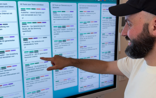 Foto: Felix Bastam steht vor einem großen Bildschirm auf dem das Trello-Board KI und Teilhabe aufgerufen ist. Er zeigt auf eine der vielen kleinen, weißen Kacheln in denen einzelne KI-Tools stehen. Felix trägt ein weißes T-Shirt und eine dunkle Kappe.
